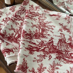 May include: Three folded linen fabric pieces with a red toile de Jouy pattern. The fabric features a cream background with a repeating design of figures in a landscape. The edges of the fabric are frayed.