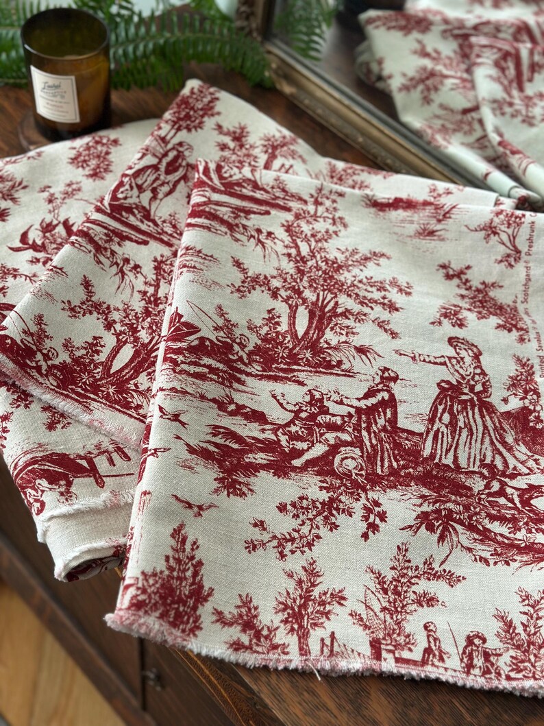 May include: Three folded linen fabric pieces with a red toile de Jouy pattern. The fabric features a cream background with a repeating design of figures in a landscape. The edges of the fabric are frayed.