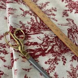 May include: Gold-handled scissors with grey blades lie on cream fabric. The fabric has a red toile de Jouy pattern depicting pastoral scenes. A wooden ruler, marked in inches, is also present on the fabric.