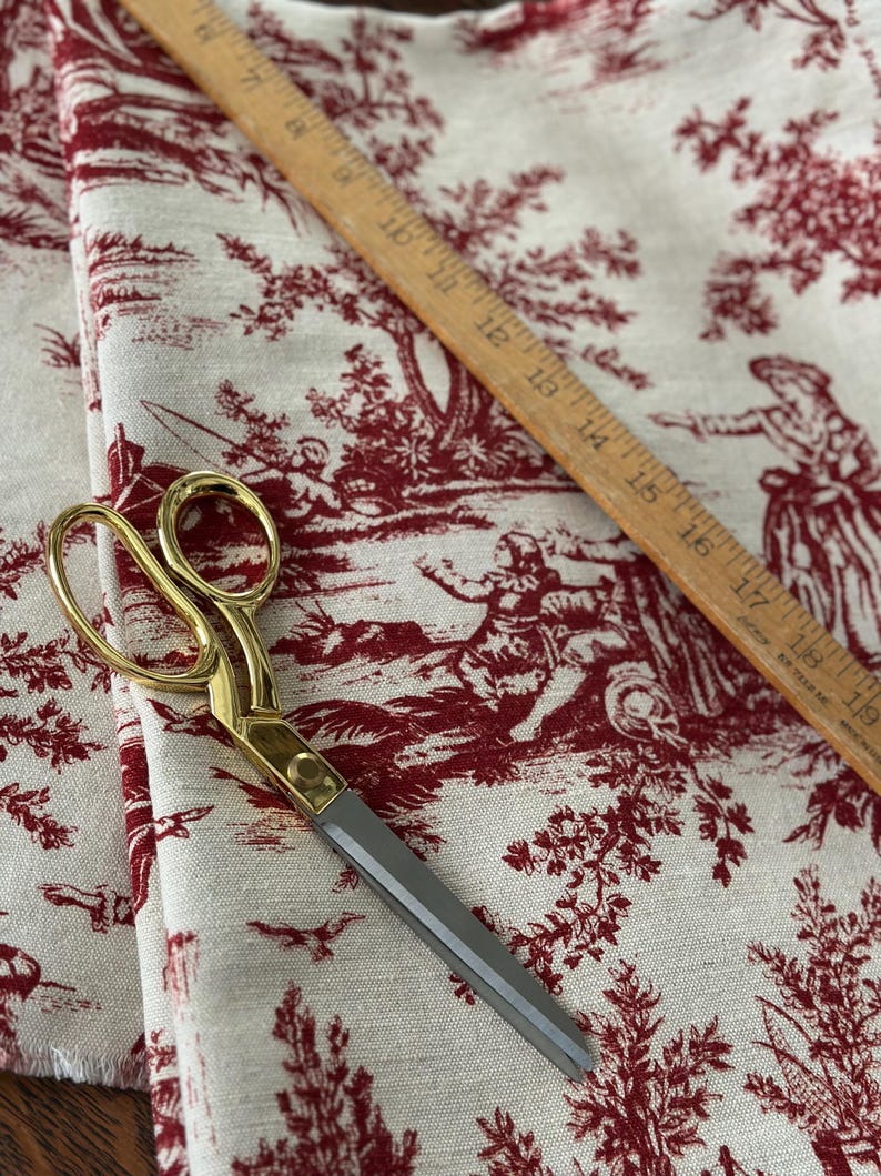 May include: Gold-handled scissors with grey blades lie on cream fabric. The fabric has a red toile de Jouy pattern depicting pastoral scenes. A wooden ruler, marked in inches, is also present on the fabric.