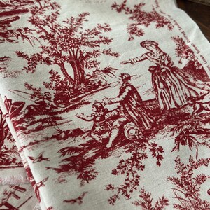 May include: Cream-coloured fabric featuring a red toile de Jouy design. The pattern shows figures in a rural setting with trees, birds, and buildings. The fabric has a raw edge, indicating it's cut from a larger piece.