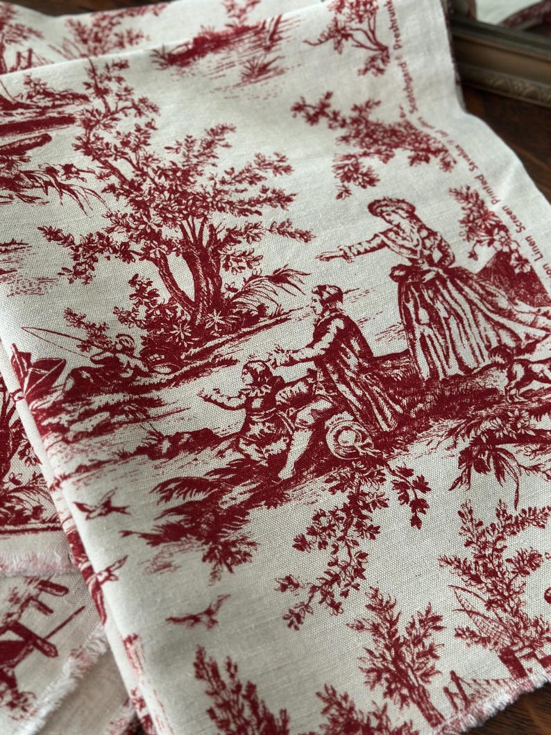 May include: Cream-coloured fabric featuring a red toile de Jouy design. The pattern shows figures in a rural setting with trees, birds, and buildings. The fabric has a raw edge, indicating it's cut from a larger piece.