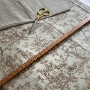 May include: A collection of sewing supplies including a beige toile fabric with a pastoral scene, a folded beige fabric, gold scissors, and a wooden ruler. The fabric features a brown and white design with figures, trees, and buildings.