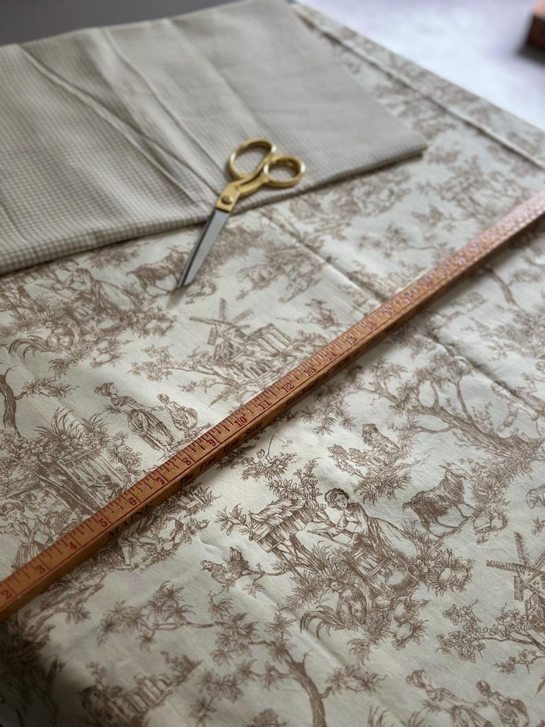 May include: A collection of sewing supplies including a beige toile fabric with a pastoral scene, a folded beige fabric, gold scissors, and a wooden ruler. The fabric features a brown and white design with figures, trees, and buildings.
