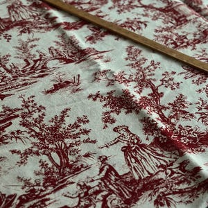 May include: Cream fabric with a red toile de Jouy design depicting rural scenes and figures. A wooden ruler lies across the fabric. The fabric is folded, displaying the intricate pattern.