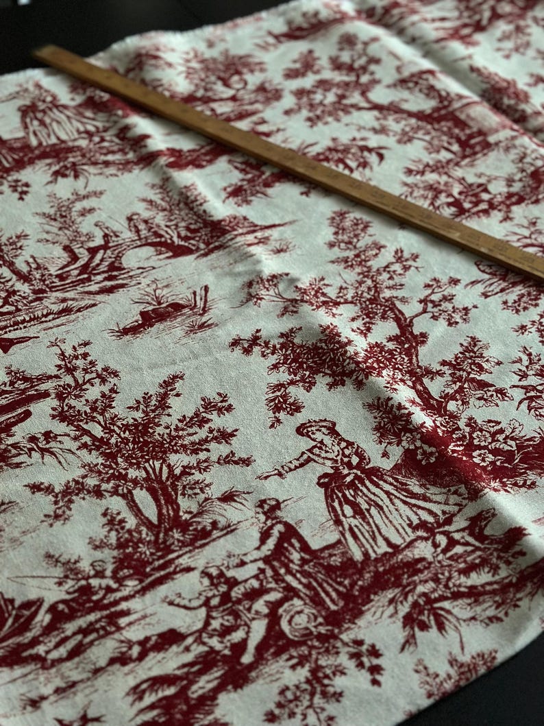 May include: Cream fabric with a red toile de Jouy design depicting rural scenes and figures. A wooden ruler lies across the fabric. The fabric is folded, displaying the intricate pattern.