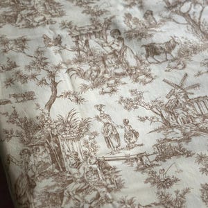 May include: Cream-colored fabric with a brown toile pattern depicting pastoral scenes. The design includes figures, buildings, trees, and animals, creating a detailed and classic aesthetic. The fabric is likely used for home decor or apparel.