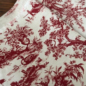 May include: Cream-coloured fabric with a repeating red toile pattern. The design features detailed illustrations of trees, ducks, and figures in a pastoral scene. The fabric is folded, showing the pattern's continuity.