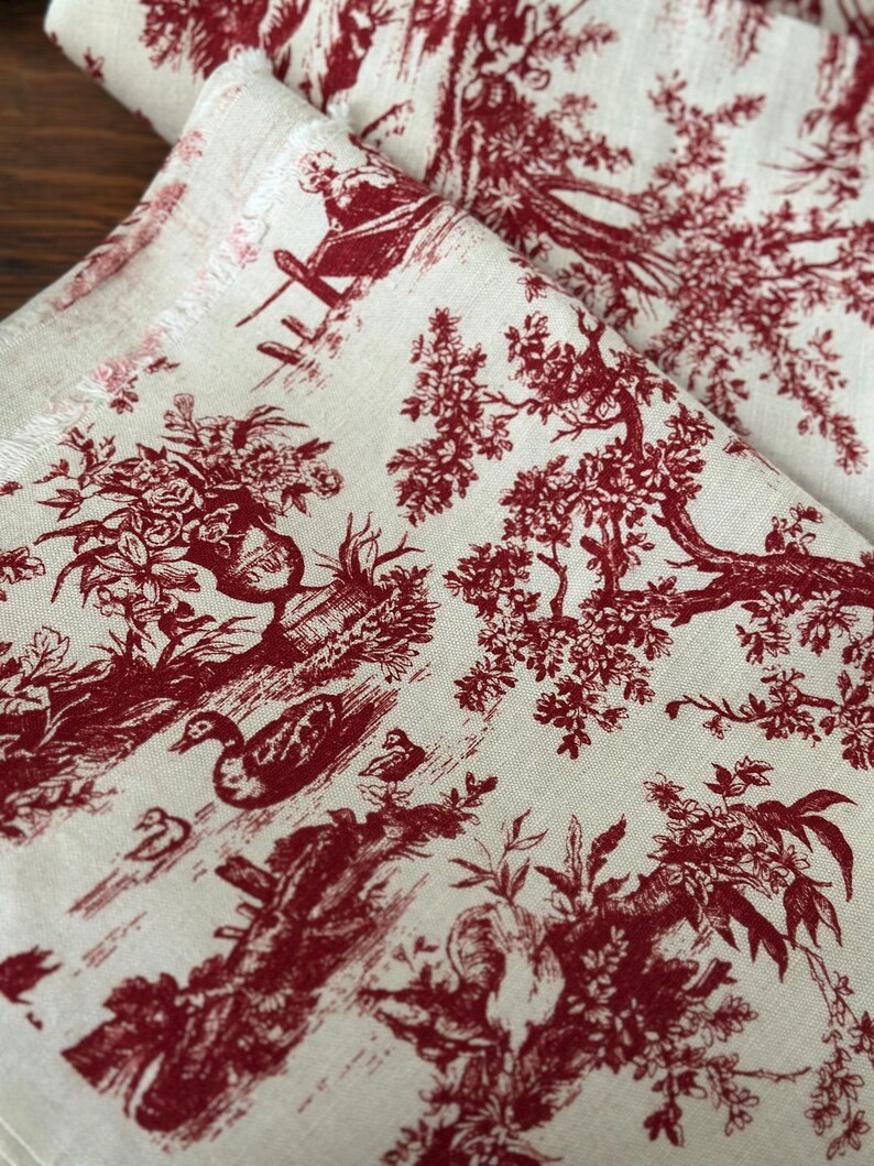May include: Cream-coloured fabric with a repeating red toile pattern. The design features detailed illustrations of trees, ducks, and figures in a pastoral scene. The fabric is folded, showing the pattern's continuity.