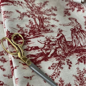 May include: A pair of gold-handled scissors lies on fabric with a red and white toile de Jouy pattern. The fabric depicts a rural scene with figures and trees. The scissors have silver blades and a gold handle.
