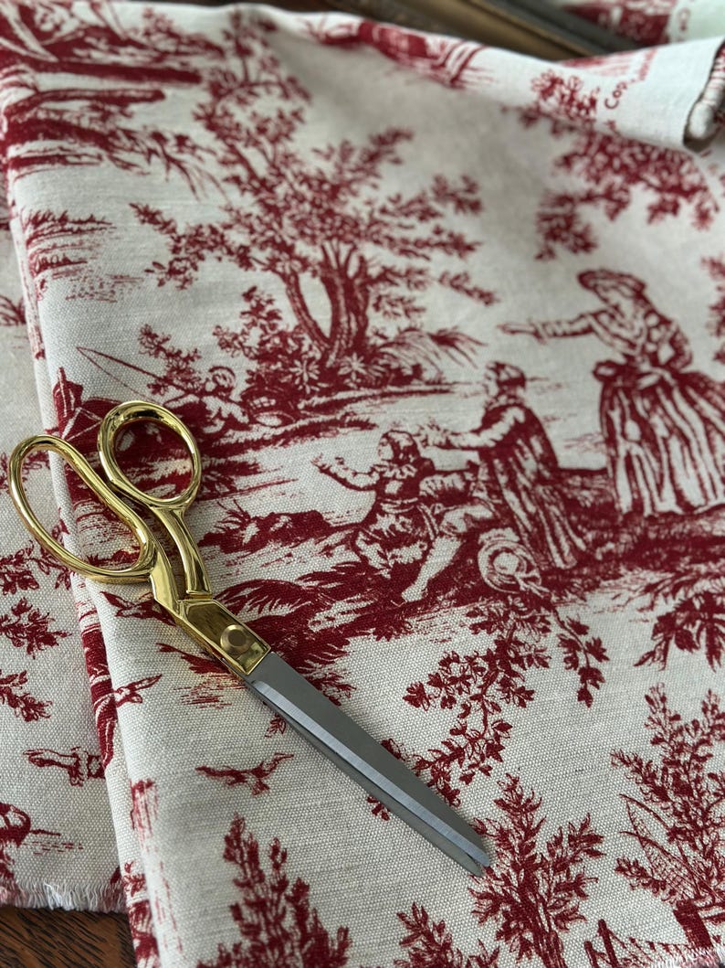 May include: A pair of gold-handled scissors lies on fabric with a red and white toile de Jouy pattern. The fabric depicts a rural scene with figures and trees. The scissors have silver blades and a gold handle.