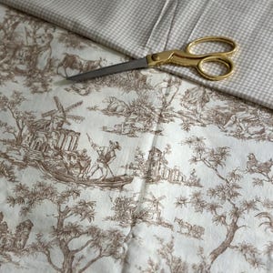 May include: A close-up of fabric and scissors. The fabric features a brown toile de Jouy pattern on a cream background. Gold scissors are positioned on top of the fabric, with a second fabric with a small checkered pattern.
