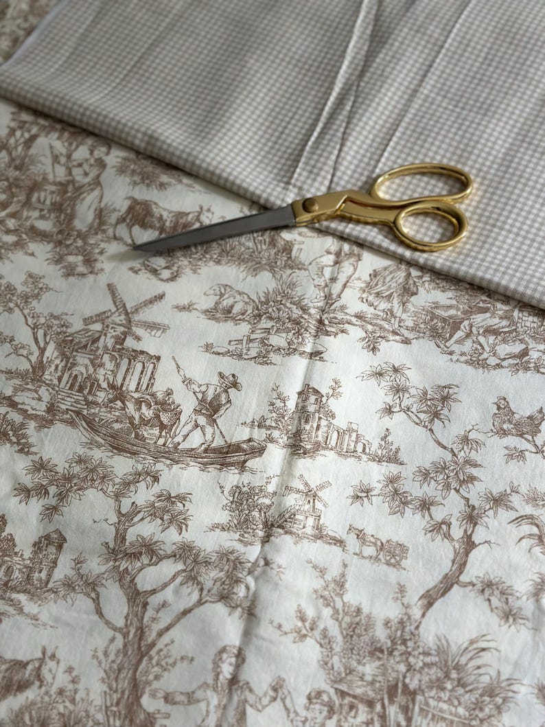 May include: A close-up of fabric and scissors. The fabric features a brown toile de Jouy pattern on a cream background. Gold scissors are positioned on top of the fabric, with a second fabric with a small checkered pattern.