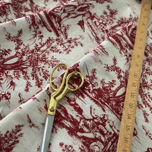 May include: Cream fabric with a red toile design depicting rural scenes. A pair of gold-handled scissors and a wooden ruler are on the fabric. The ruler measures up to 45 cm.