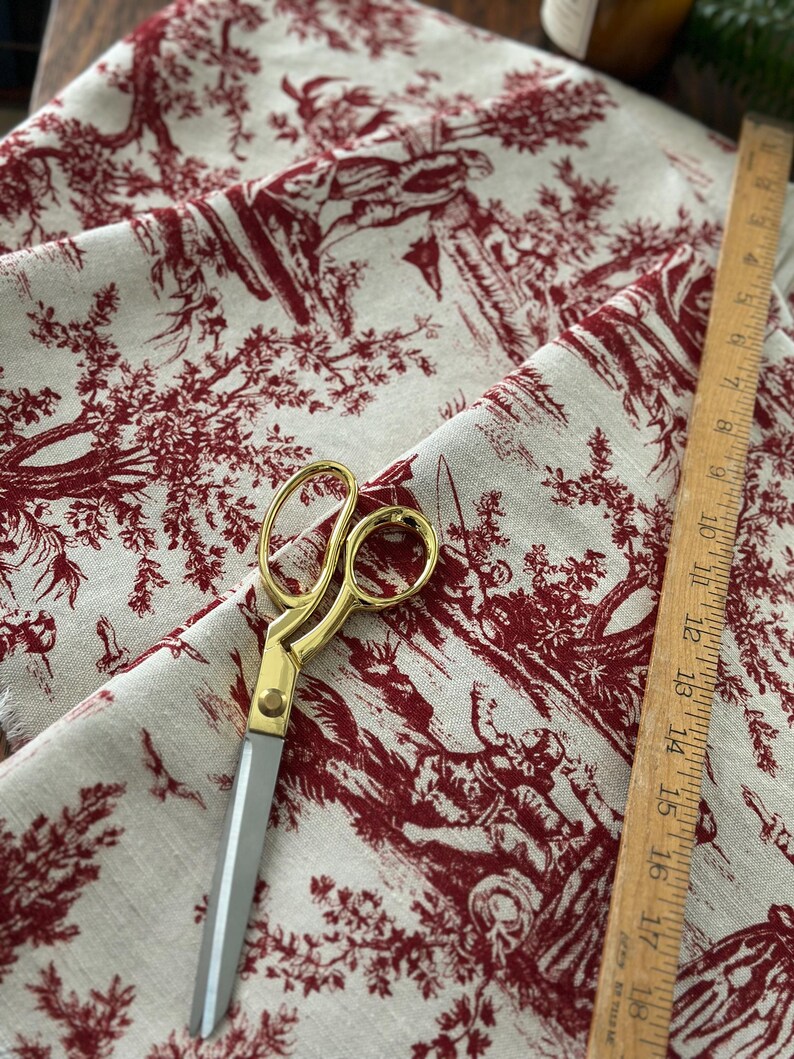 May include: Cream fabric with a red toile design depicting rural scenes. A pair of gold-handled scissors and a wooden ruler are on the fabric. The ruler measures up to 45 cm.