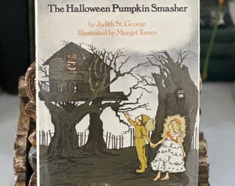 以下が含まれることがあります： ジュディス・セント・ジョージ著、マーゴット・トームズ画のハードカバー本『The Halloween Pumpkin Smasher』。表紙には、窓に光るジャック・オー・ランタンが飾られた暗いツリーハウスと、その下に立つ仮装した二人の子供が描かれています。