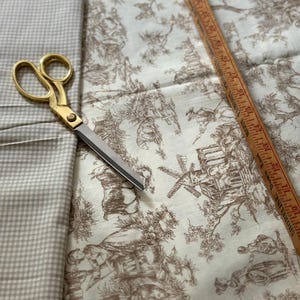 May include: A craft scene featuring fabric, scissors, and a ruler. The fabric includes a beige gingham pattern and a toile design with brown illustrations on a cream background. Gold scissors and a wooden ruler are also present.