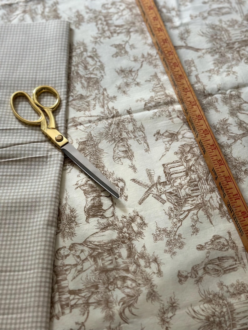 May include: A craft scene featuring fabric, scissors, and a ruler. The fabric includes a beige gingham pattern and a toile design with brown illustrations on a cream background. Gold scissors and a wooden ruler are also present.