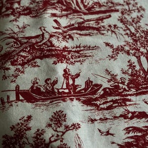 May include: Close-up of fabric with a red toile de Jouy pattern. The design features scenes of people in boats, birds, trees, and fountains against a cream background. The fabric has a classic, vintage aesthetic.