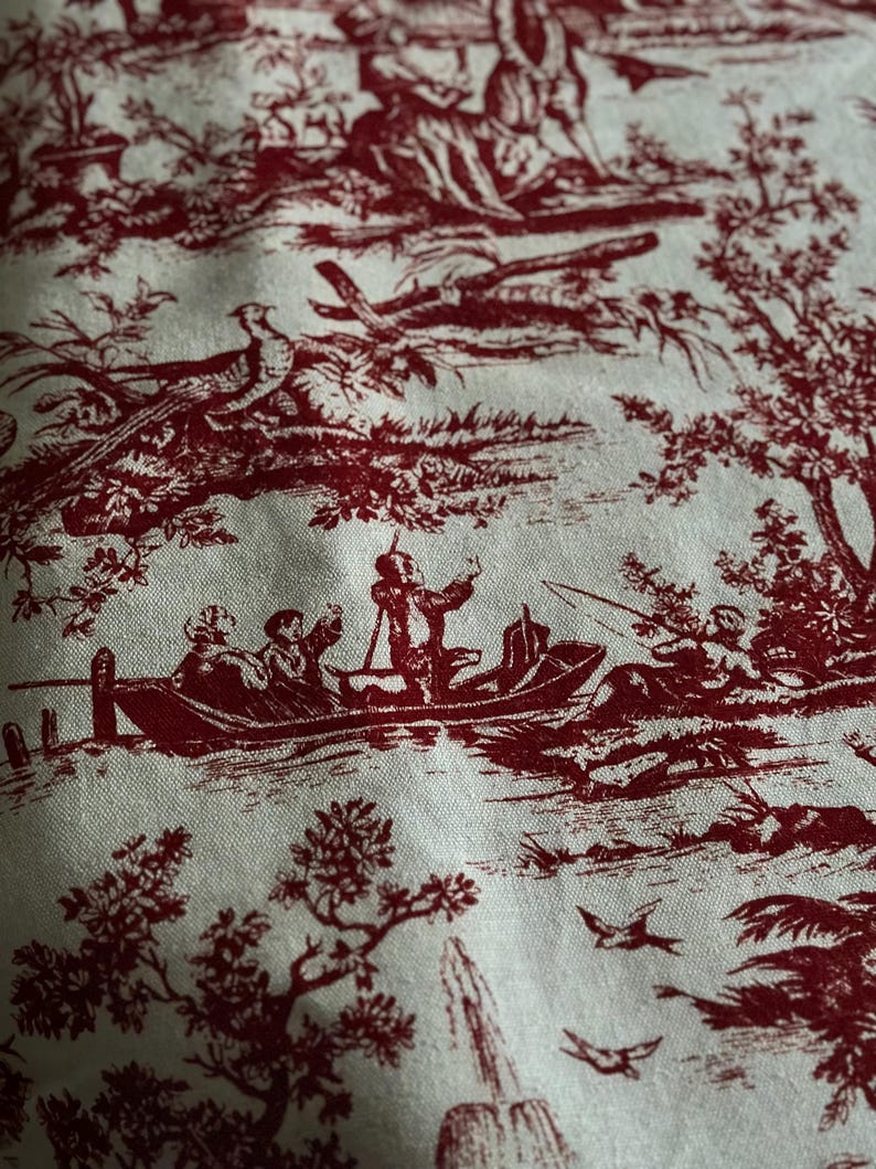 May include: Close-up of fabric with a red toile de Jouy pattern. The design features scenes of people in boats, birds, trees, and fountains against a cream background. The fabric has a classic, vintage aesthetic.