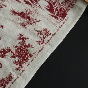 May include: Close-up of linen fabric with a red toile de Jouy design. The fabric depicts a pastoral scene with figures, trees, and a bridge. The text "Linen Screen Printed Savalux fast colors Scotchgard Preshrunk Copyright Gabrielle Cie" is printed on the fabric.