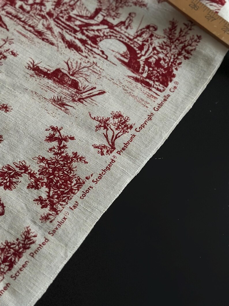 May include: Close-up of linen fabric with a red toile de Jouy design. The fabric depicts a pastoral scene with figures, trees, and a bridge. The text "Linen Screen Printed Savalux fast colors Scotchgard Preshrunk Copyright Gabrielle Cie" is printed on the fabric.