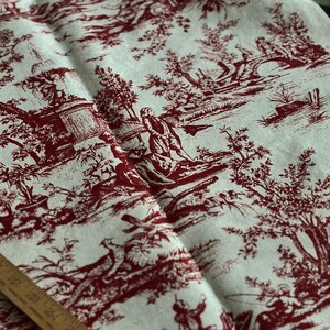 May include: Cream fabric featuring a burgundy toile de Jouy design. The pattern shows scenes of people in a landscape with trees, bridges, and figures. A wooden ruler is visible along the bottom edge of the fabric.