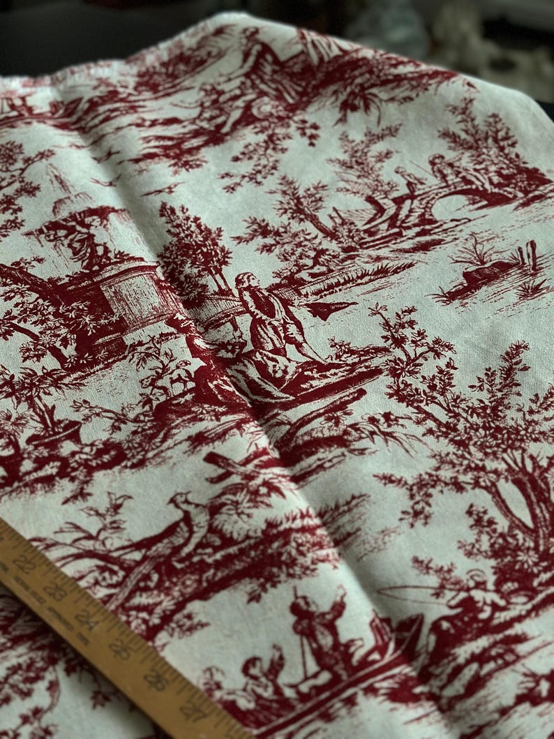 May include: Cream fabric featuring a burgundy toile de Jouy design. The pattern shows scenes of people in a landscape with trees, bridges, and figures. A wooden ruler is visible along the bottom edge of the fabric.
