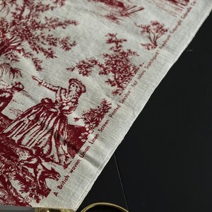 May include: A piece of fabric with a red and white toile de Jouy design, depicting scenes of people and nature. The fabric has the text "A British woven Union Linen Screen Printed Savalux fast colors Scotchgard Preshrunk Copyright Gabrielle C". Gold scissors are visible.