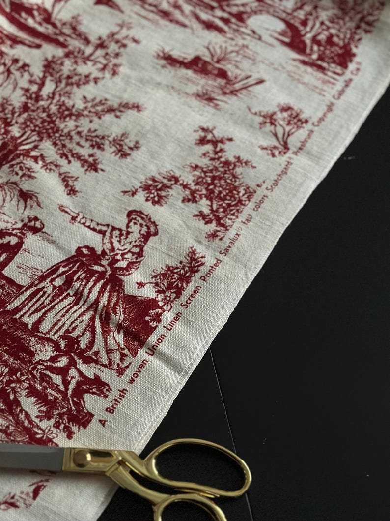May include: A piece of fabric with a red and white toile de Jouy design, depicting scenes of people and nature. The fabric has the text "A British woven Union Linen Screen Printed Savalux fast colors Scotchgard Preshrunk Copyright Gabrielle C". Gold scissors are visible.