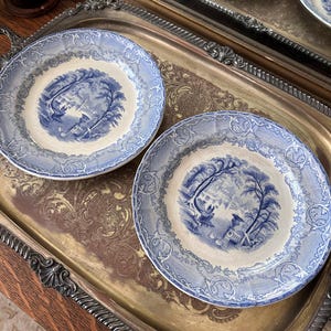 May include: Two blue and white porcelain plates with a scenic design, resting on a decorative silver tray. The plates feature a detailed landscape scene in the center and a patterned border. The tray has ornate handles and a detailed embossed design.