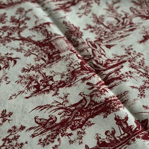 May include: Folded fabric with a traditional toile de Jouy design. The burgundy pattern on a cream backdrop shows scenes of figures, trees, and boats. The fabric, likely linen or a similar material, is folded, revealing the detailed print.