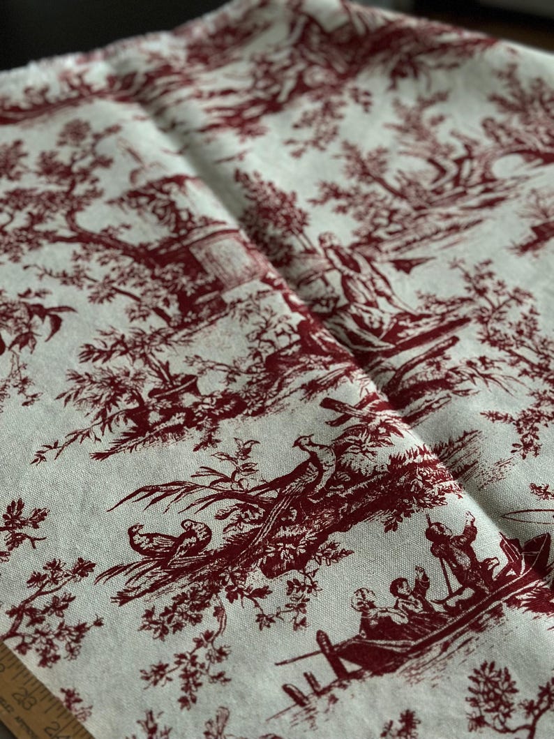 May include: Folded fabric with a traditional toile de Jouy design. The burgundy pattern on a cream backdrop shows scenes of figures, trees, and boats. The fabric, likely linen or a similar material, is folded, revealing the detailed print.