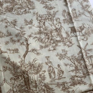May include: Cream-colored fabric with a brown toile de Jouy pattern. The design depicts pastoral scenes with figures, animals, and buildings. The fabric is folded, showing the detailed print.