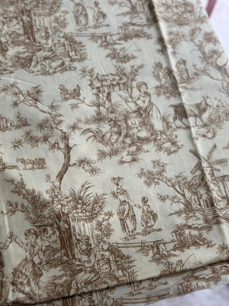 May include: Cream-colored fabric with a brown toile de Jouy pattern. The design depicts pastoral scenes with figures, animals, and buildings. The fabric is folded, showing the detailed print.