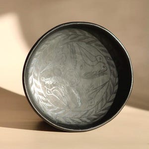 May include: A dark gray, round metal bowl with an etched design of birds and foliage. The bowl has a slightly raised rim and a matte finish. The interior design is in a lighter gray color.