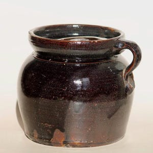 May include: A dark brown, glazed ceramic jar with a rounded body and a single handle. The jar has a wide opening and a slightly flared rim. The surface has a speckled appearance, and the jar appears to be vintage or antique.