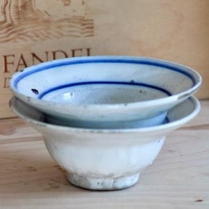 May include: A small, white ceramic bowl with a flared rim and a footed base. The interior of the bowl features two distinct, horizontal blue painted lines circling the inner wall. The exterior surface is plain white with visible signs of wear.