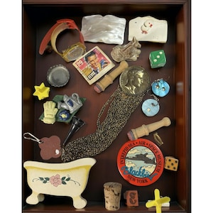 Puede incluir: Una colección de artículos vintage que incluye un reloj, un sello de Elvis, dados, una figura de bañera y un pin del Museo Espacial de Nueva York, dispuestos en una caja de madera. Los artículos son de varios colores y materiales.