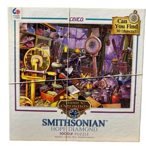May include: A Smithsonian Hope Diamond 1000-piece puzzle. The box shows a detailed illustration of a cluttered room with various objects. The puzzle is labeled "Hidden Expedition" and "Can You Find 30 Objects?" Made in the USA.