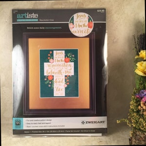 Puede incluir: Un kit de punto de cruz contado con una cruz verde adornada con flores y el texto "Jesús, yo soy la resurrección y la vida, el que cree en mí, aunque esté muerto, vivirá." El kit es de la marca Artiste y se llama "Resurrection Cross".