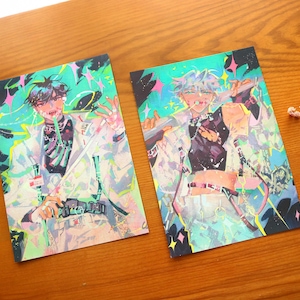 May include: Two art prints with anime-style illustrations of figures in white coats and black tops. The artwork has a vibrant colour palette with accents of green, pink, and blue. The prints are displayed on a wooden surface.