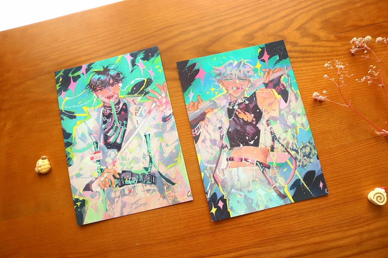 May include: Two art prints with anime-style illustrations of figures in white coats and black tops. The artwork has a vibrant colour palette with accents of green, pink, and blue. The prints are displayed on a wooden surface.