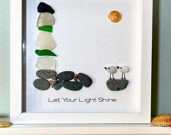May include: A white framed picture featuring a sea glass and stone artwork. The artwork depicts a lighthouse, a sun, and two birds perched on a rock. The text "Let Your Light Shine" is written beneath the artwork.