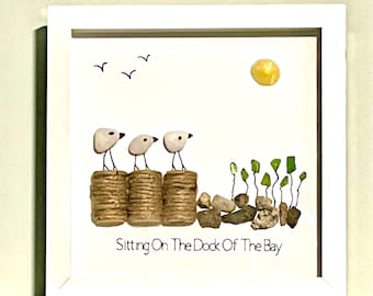 May include: A framed pebble art picture with three white birds perched on a wooden dock. The background is a light blue sky with a yellow sun and three white birds flying. The text "Sitting On The Dock Of The Bay" is written below the birds.