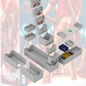 Puede incluir: Una caja de cartón blanca con divisores para organizar un juego de mesa. La caja tiene compartimentos etiquetados como "Eventos", "Configuración del jugador" y "Conciertos aleatorios". La caja está diseñada para contener los componentes del juego de mesa "Rock Hard - 1977".