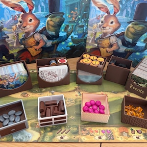 May include: A game board with various wooden storage boxes, including a box with the word "Events" on it. The boxes contain game pieces, cards, and tokens. The board is decorated with a whimsical illustration of a rabbit and a turtle.