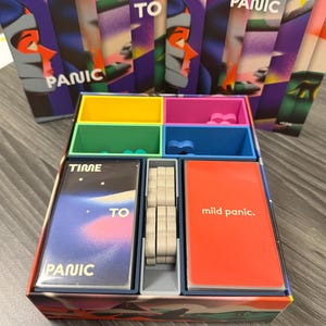 Puede incluir: Un juego de mesa llamado "Time to Panic" con una caja y cartas. La caja está dividida en compartimentos de colores. Las cartas tienen el título del juego y las palabras "mild panic". El fondo presenta cartas con el perfil de una persona.