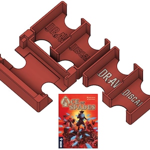 May include: Red card game organizers with the words "DRAW" and "DISCARD" printed on them. The organizers are next to a game box titled "ACE OF SPADES" with a colorful illustration of characters.