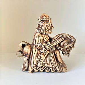 Puede incluir: Figura beige de un rey a caballo. El rey lleva una corona y empuña una espada. El caballo está tallado con intrincados detalles. La pieza tiene un aspecto vintage, con detallados relieves y color crema.